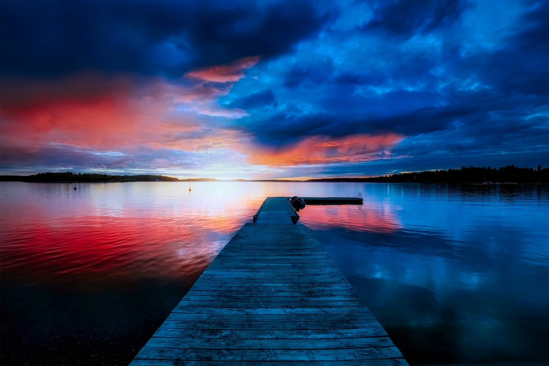 A tranquil sunset view with vibrant hues reflecting over a calm lake and a wooden dock extending into the water.