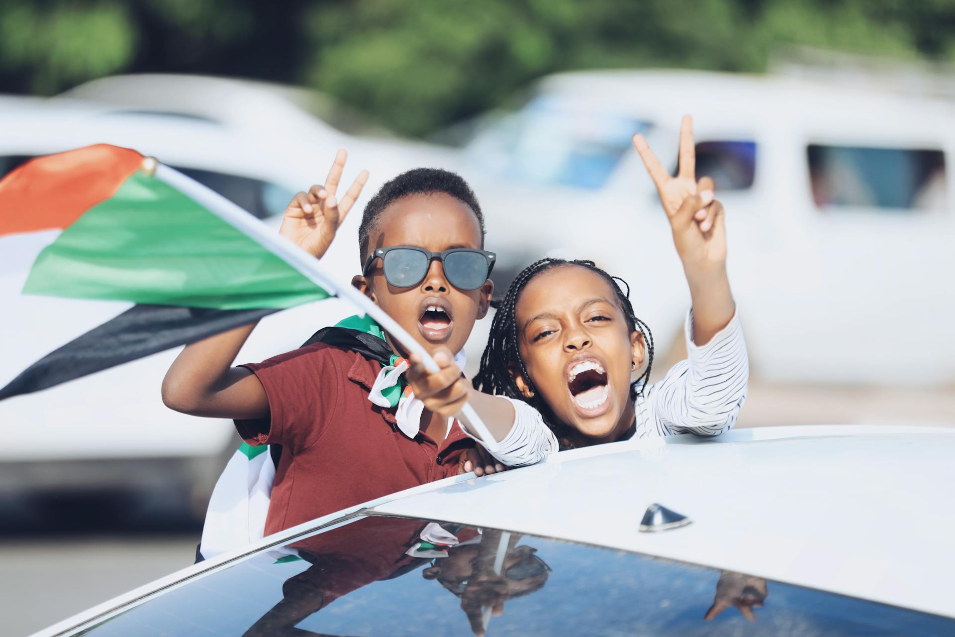 Energetic children expressing joy with peace signs, waving the Sudanese flag from a car.