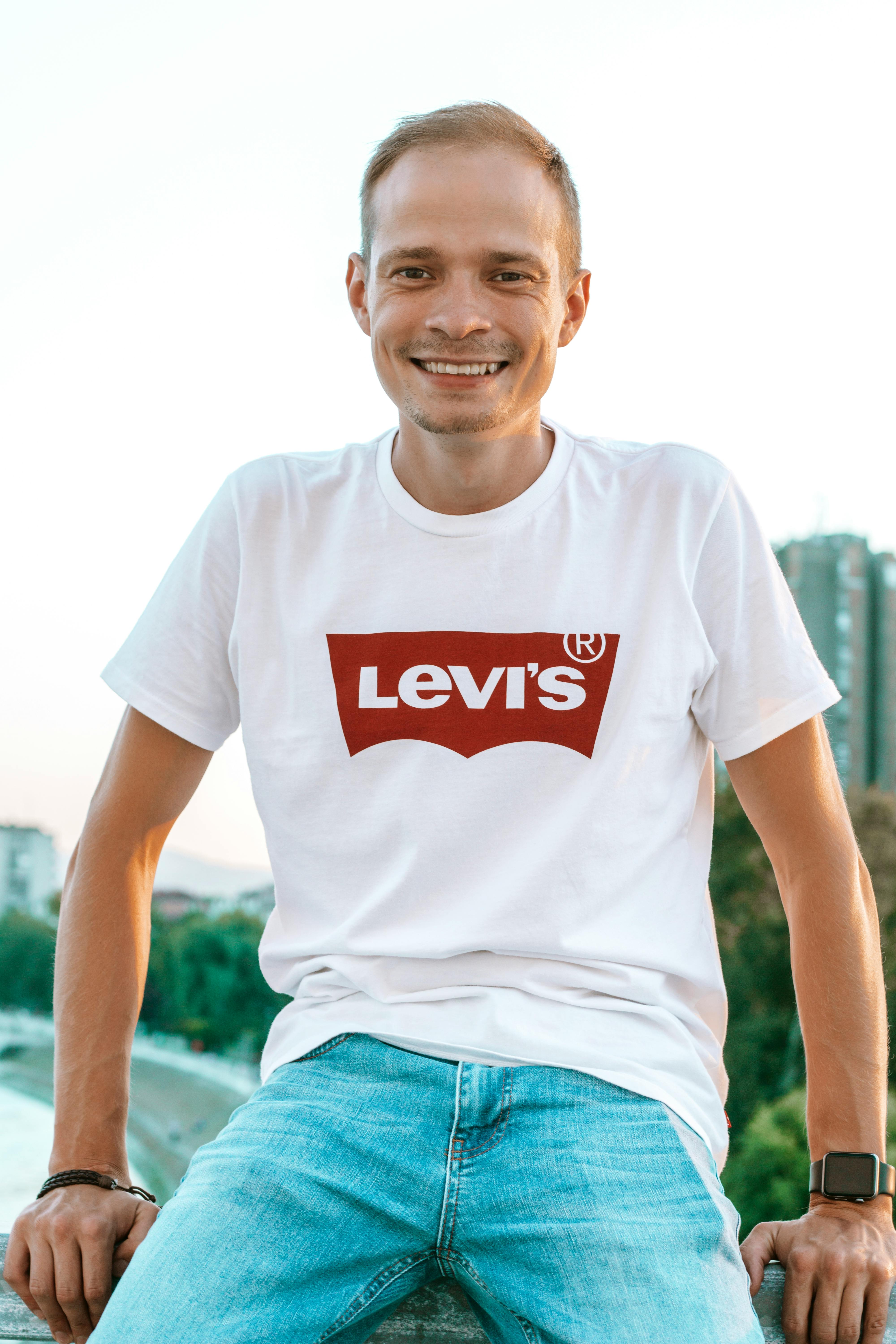 Low angle of young happy guy in trendy t shirt and smart watch sitting in town under sky and looking at camera