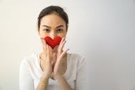 Unrecognizable female looking at camera while standing on white background with small handmade heart in hands covering mouth in room