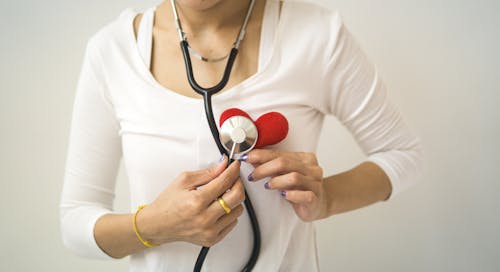 Unrecognizable female wearing white shirt while standing on white background with diaphragm of stethoscope on red handmade heart in room