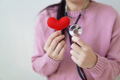Free Unrecognizable female in pink sweater with stethoscope on neck standing on white background with red heart in hand in daylight Stock Photo
