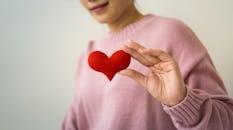 Content unrecognizable female wearing pink sweater standing on white background with small handmade heart in hand in light room inside