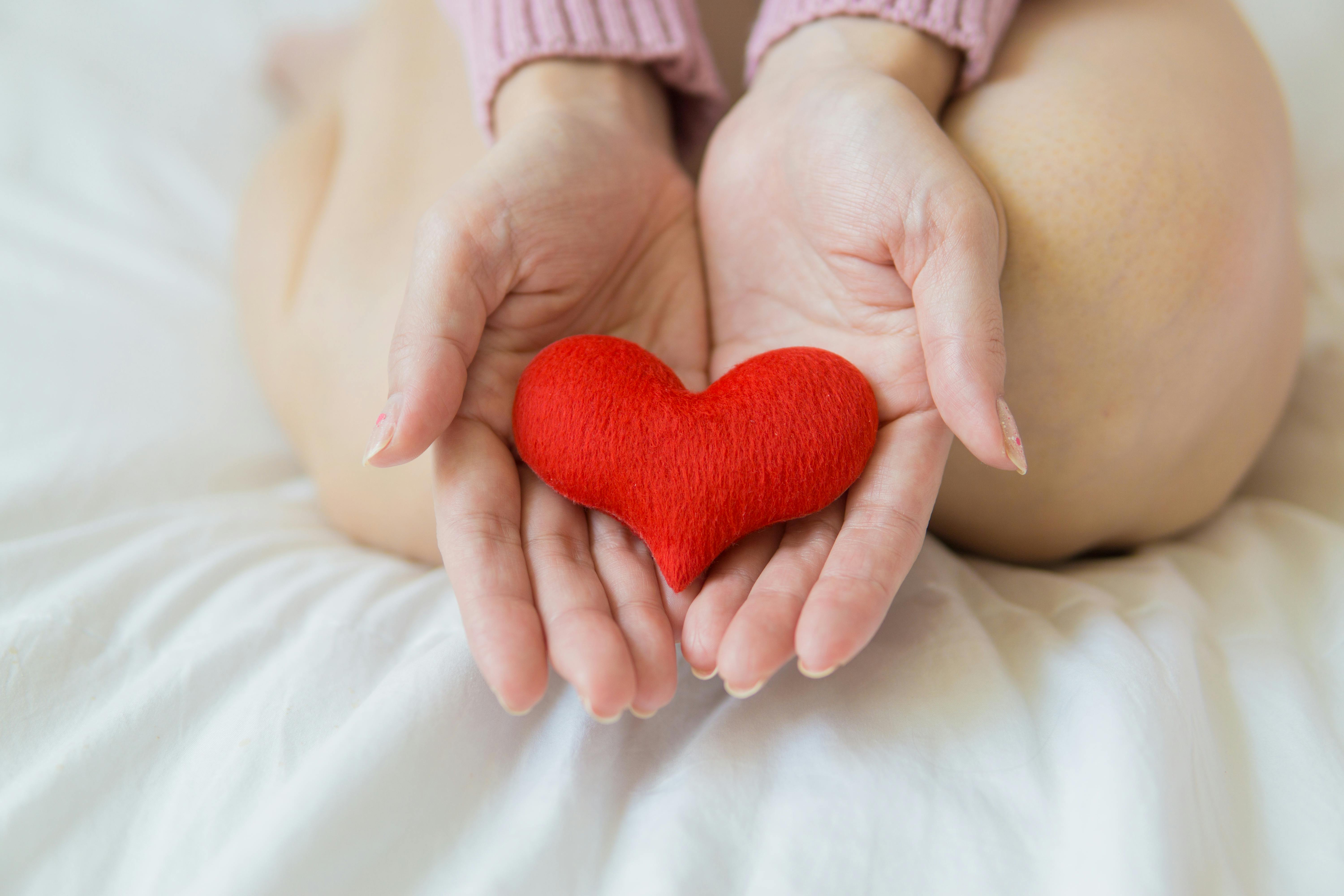 Free Unrecognizable female sitting with bare legs on white sheet with small red heart in hands in light room in daytime Stock Photo