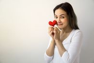 房间里，一位留着长发的快乐女性站在白色背景前，脸上带着红心，注视着镜头