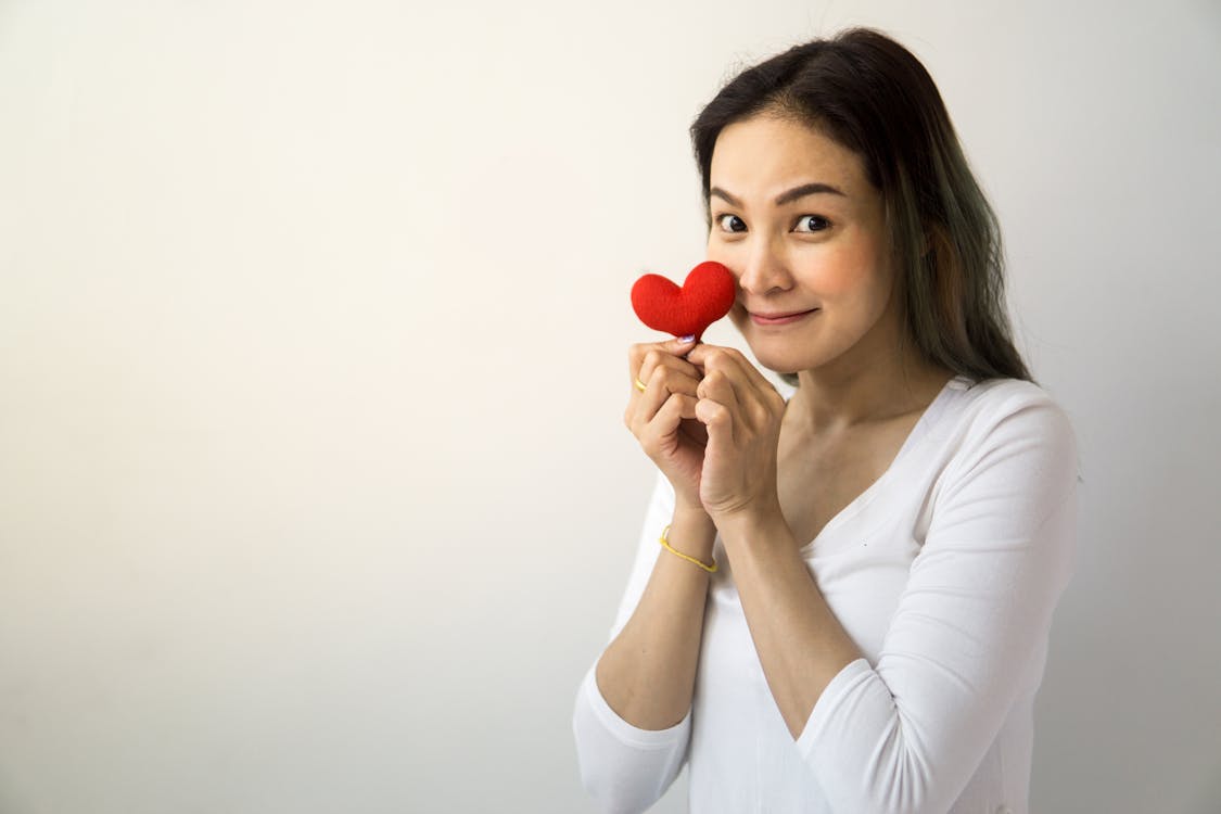 Free Cheerful female with long hair looking at camera while standing on white background with red heart near face in room Stock Photo