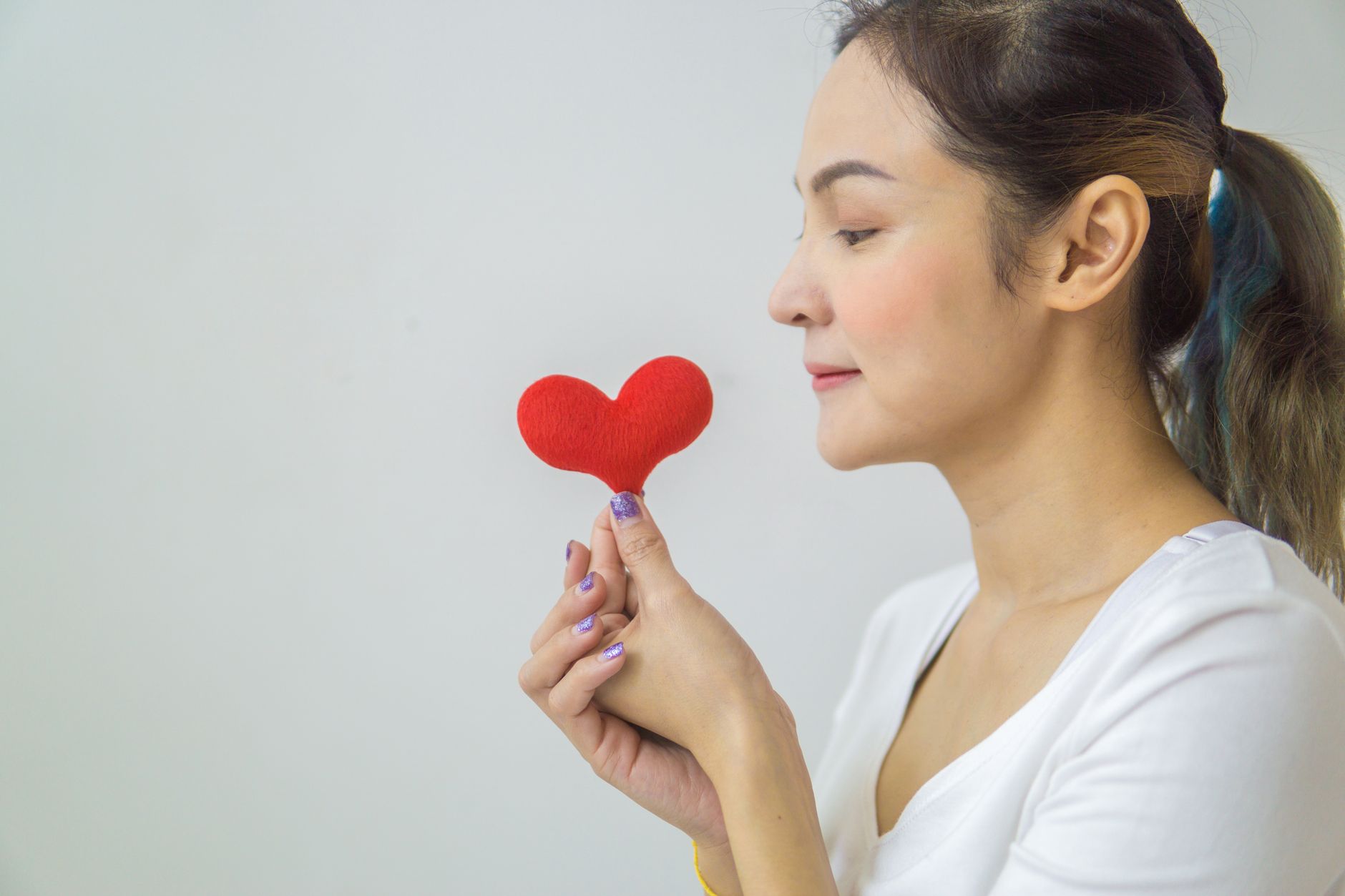 Side view of calm female in ponytail with red heart in hand and standing on white background in light room