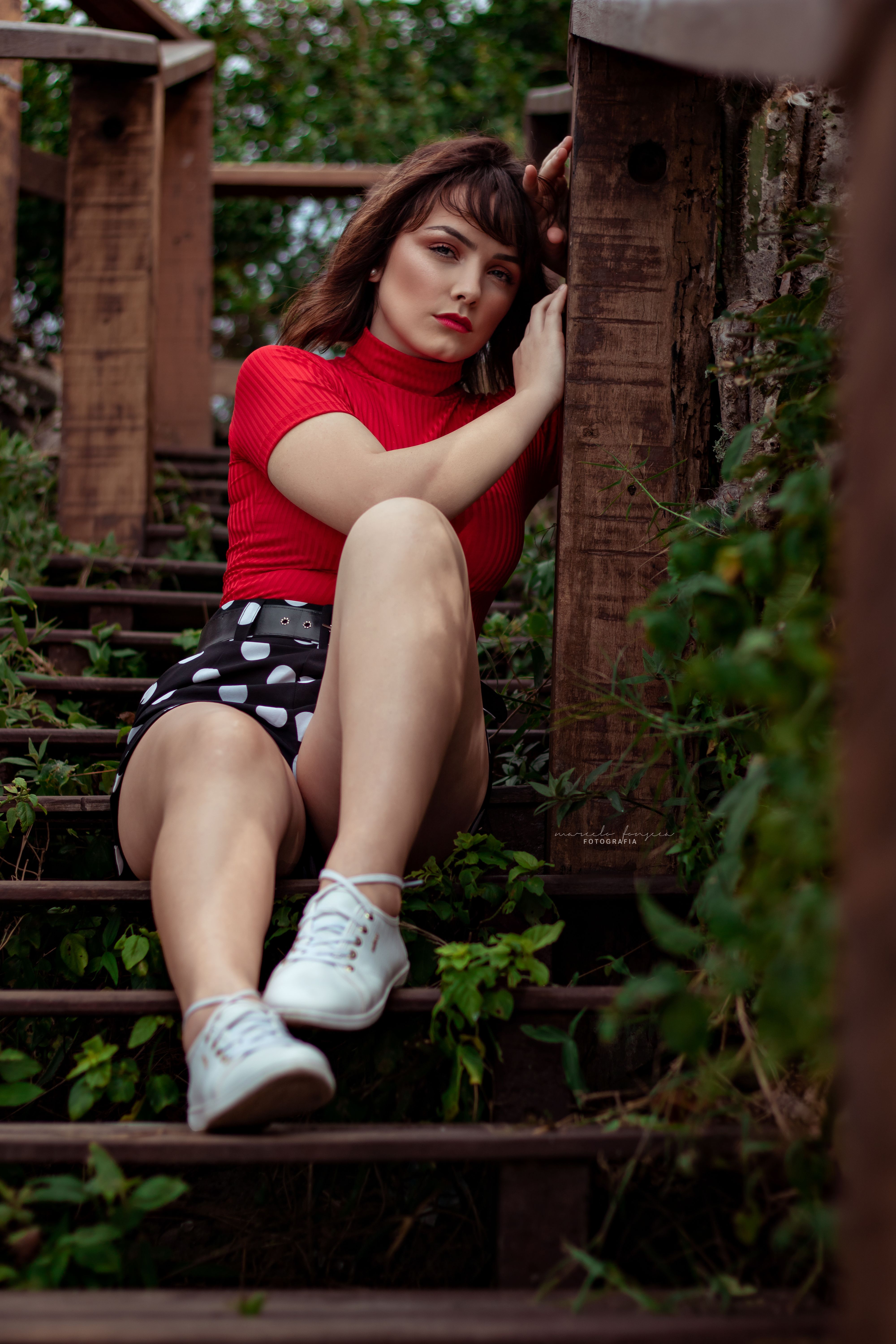 Portrait of a young woman sitting on wooden stairs surrounded by greenery in a relaxed pose.