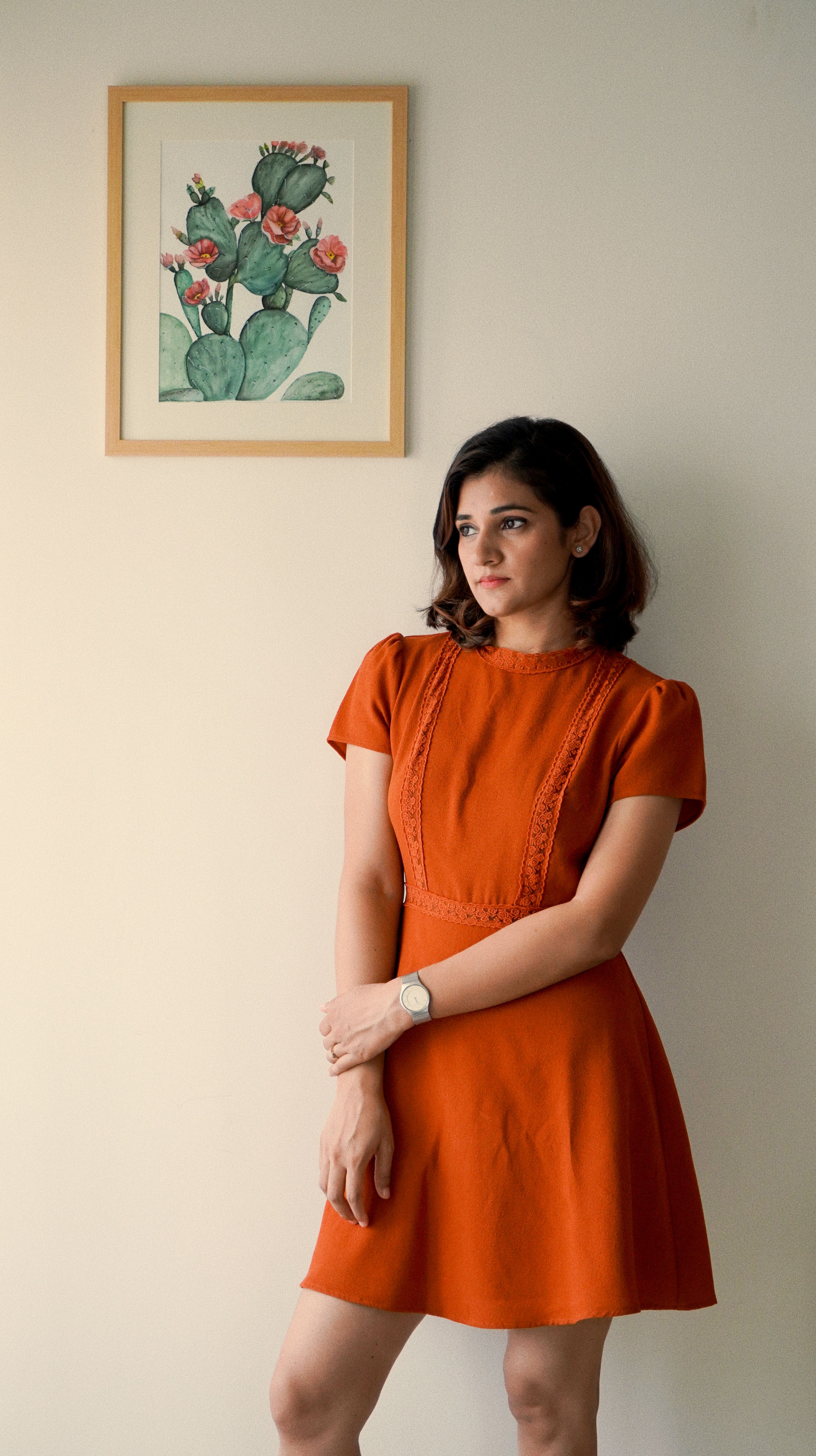 Woman in an orange dress standing indoors near a framed artwork, looking away.