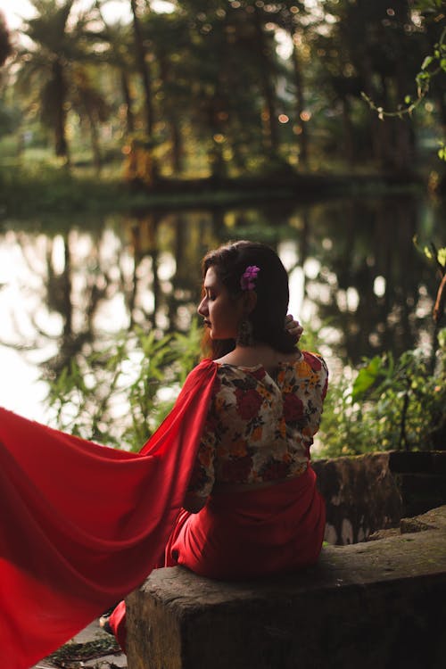 Back view of serene young Indian female in red flying sari looking down while resting on stone slap in park next to reservoir on blurred background