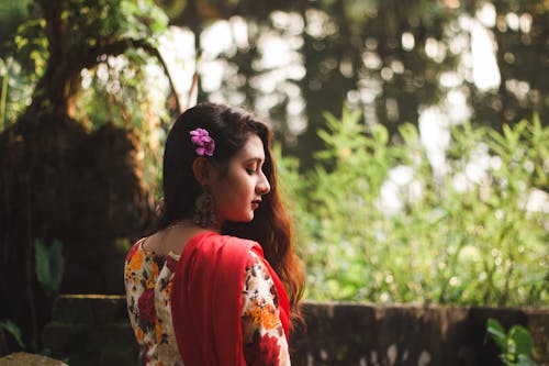 Side view of pensive young Indian female in traditional garment with small pink flower in dark wavy hair looking down while chilling in park by pond in daytime on blurred background