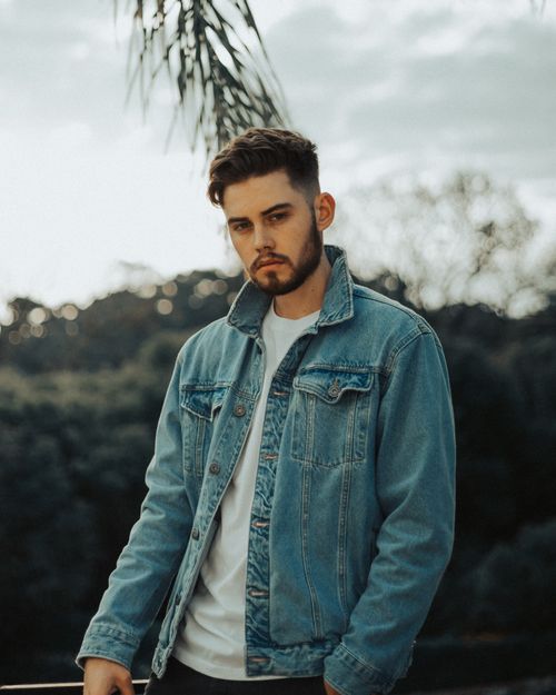 Free Young man with a beard in a denim jacket posing outdoors with a moody expression. Stock Photo