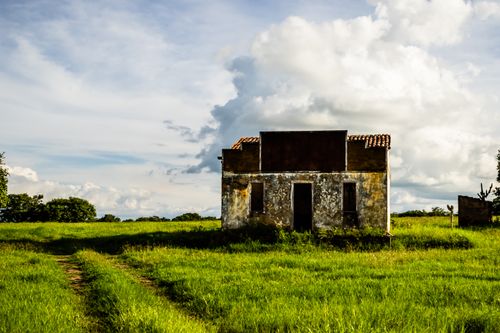 Free A rustic abandoned house stands alone in a vibrant green grass field under a cloudy sky. Stock Photo