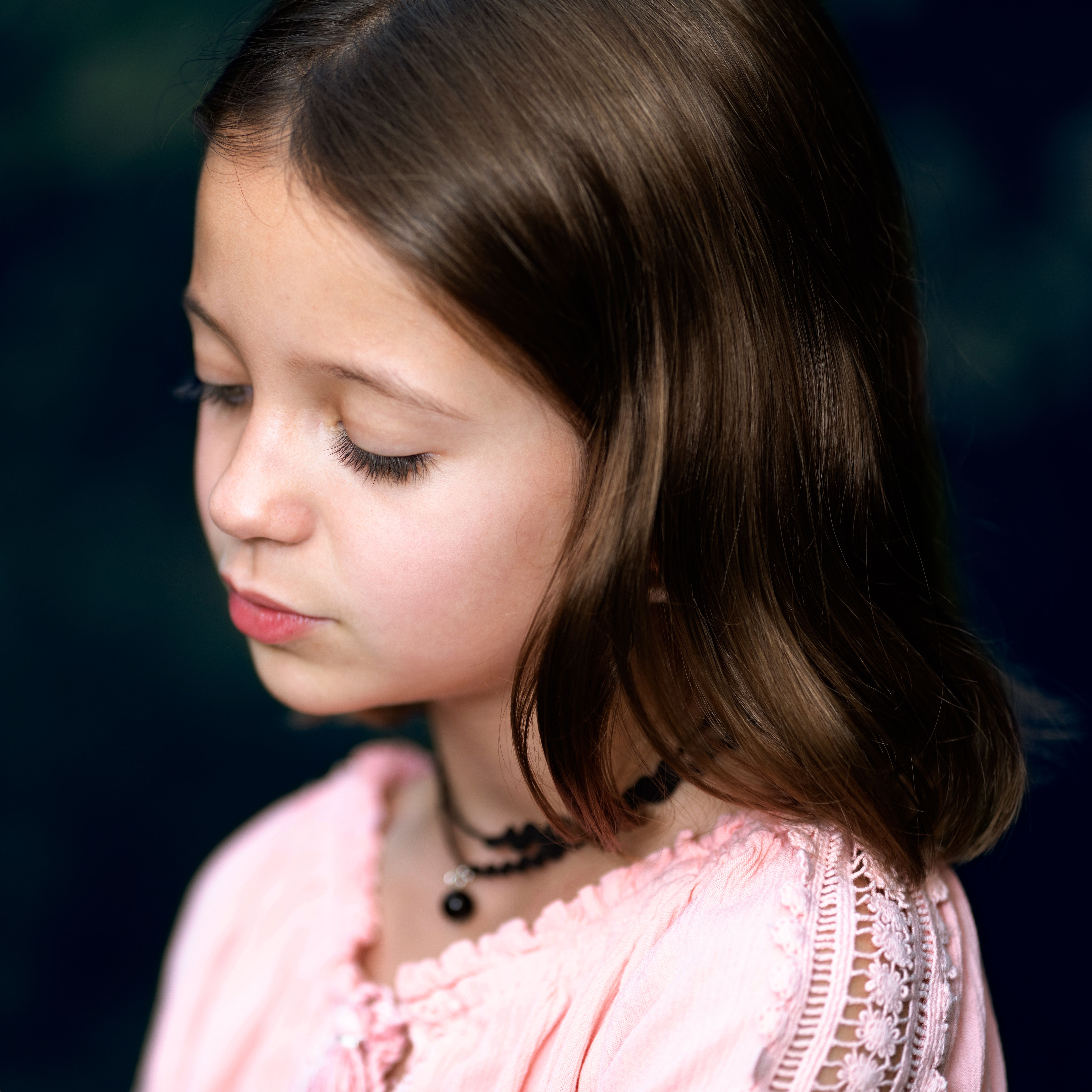 Crop wistful girl in rose wear and beads with brown hair looking down on dark background
