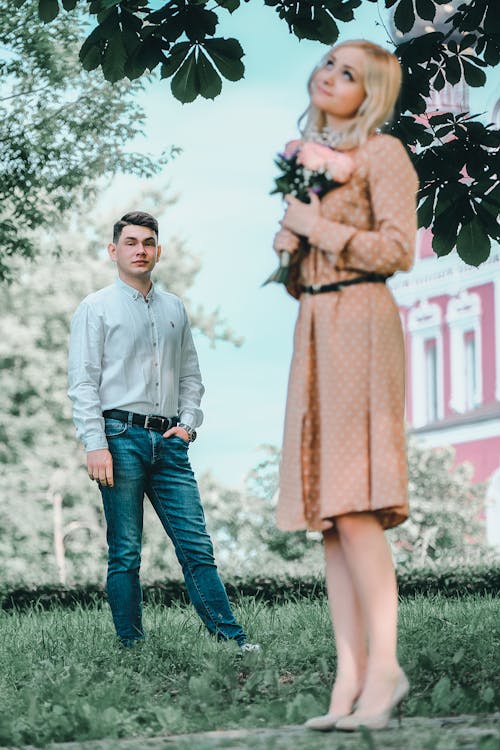 Free Full length charming female in stylish dress standing with bouquet in green park near young boyfriend against ornamental building Stock Photo