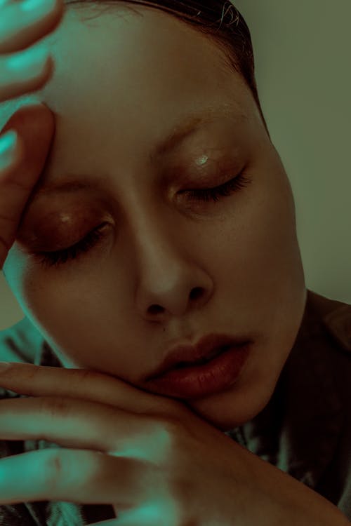 Headshot of attractive female with closed eyes and bright make up with hands near face in dim light in room