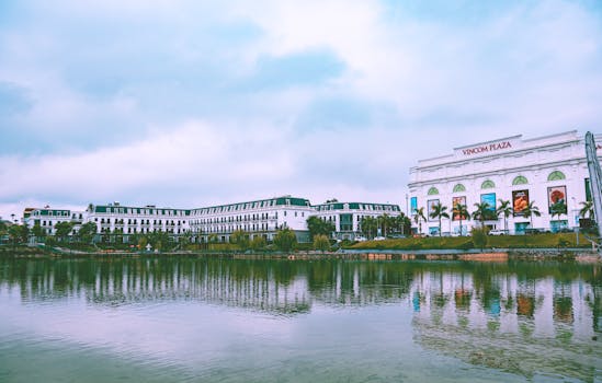 A view of Vincom Plaza and adjacent buildings reflecting off a calm lake under a cloudy sky.