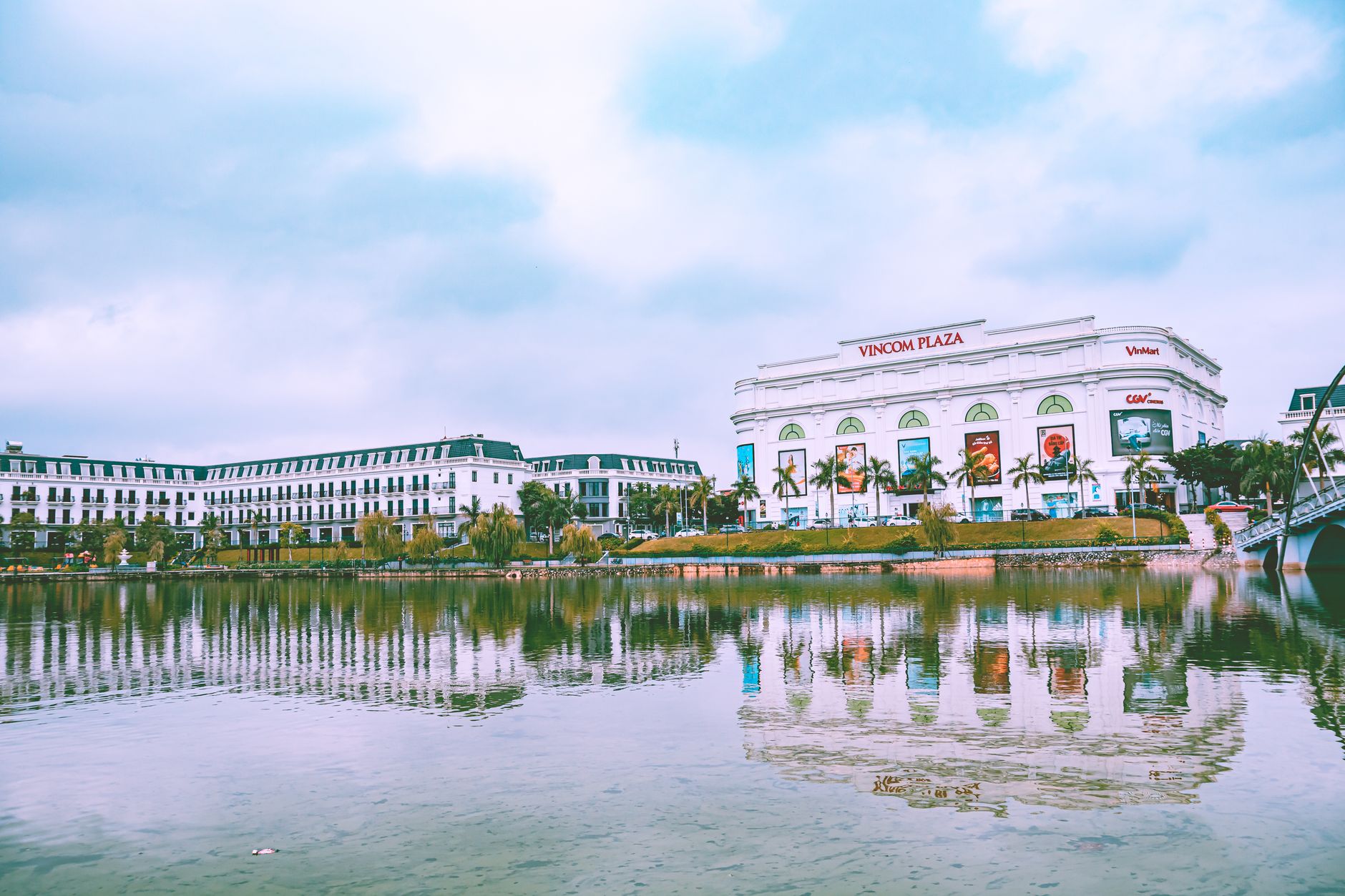 Urban cityscape featuring Vincom Plaza reflecting in a serene lake, showcasing modern architecture and palm trees.