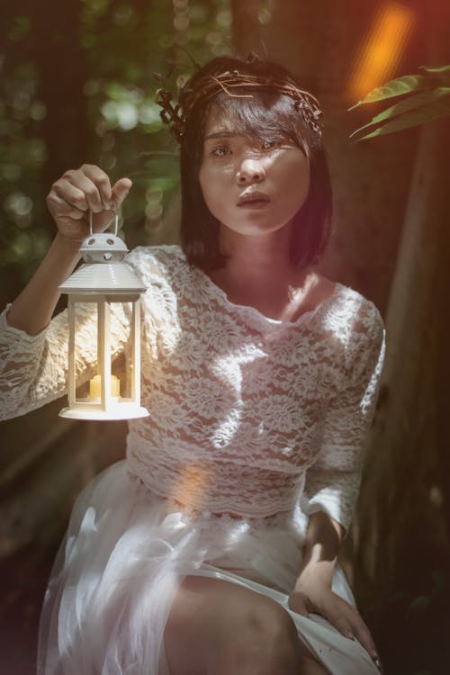 Calm Asian female in crown looking at camera while sitting in forest near green leaves under sun beams with lamp in hand