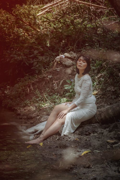Full body of barefoot Asian female with closed eyes and long dress sitting on beam near water with hand on knee