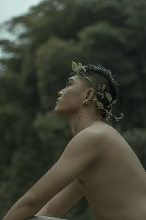 Side view of young Asian serious shirtless guy with natural wreath on hand relaxing in nature on gloomy day