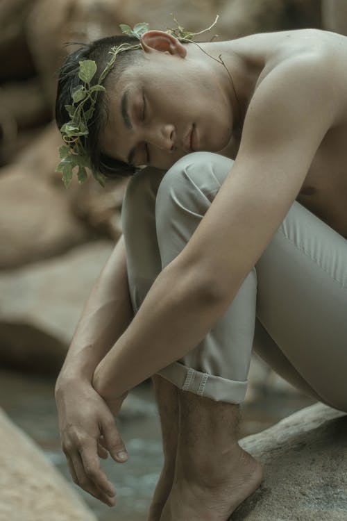 Side view of tranquil barefooted young Asian male with naked torso and leaf wreath on hand embracing knees with closed eyes while sitting on stone near river