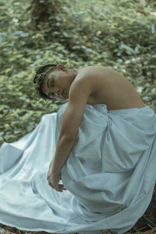 Side view of calm young Asian guy with naked torso and leaf wreath covering body with white bed sheet while relaxing in green forest