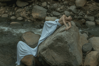 Male in white sheet and organic wreath looking at camera and lying on rough stone near foamy stream