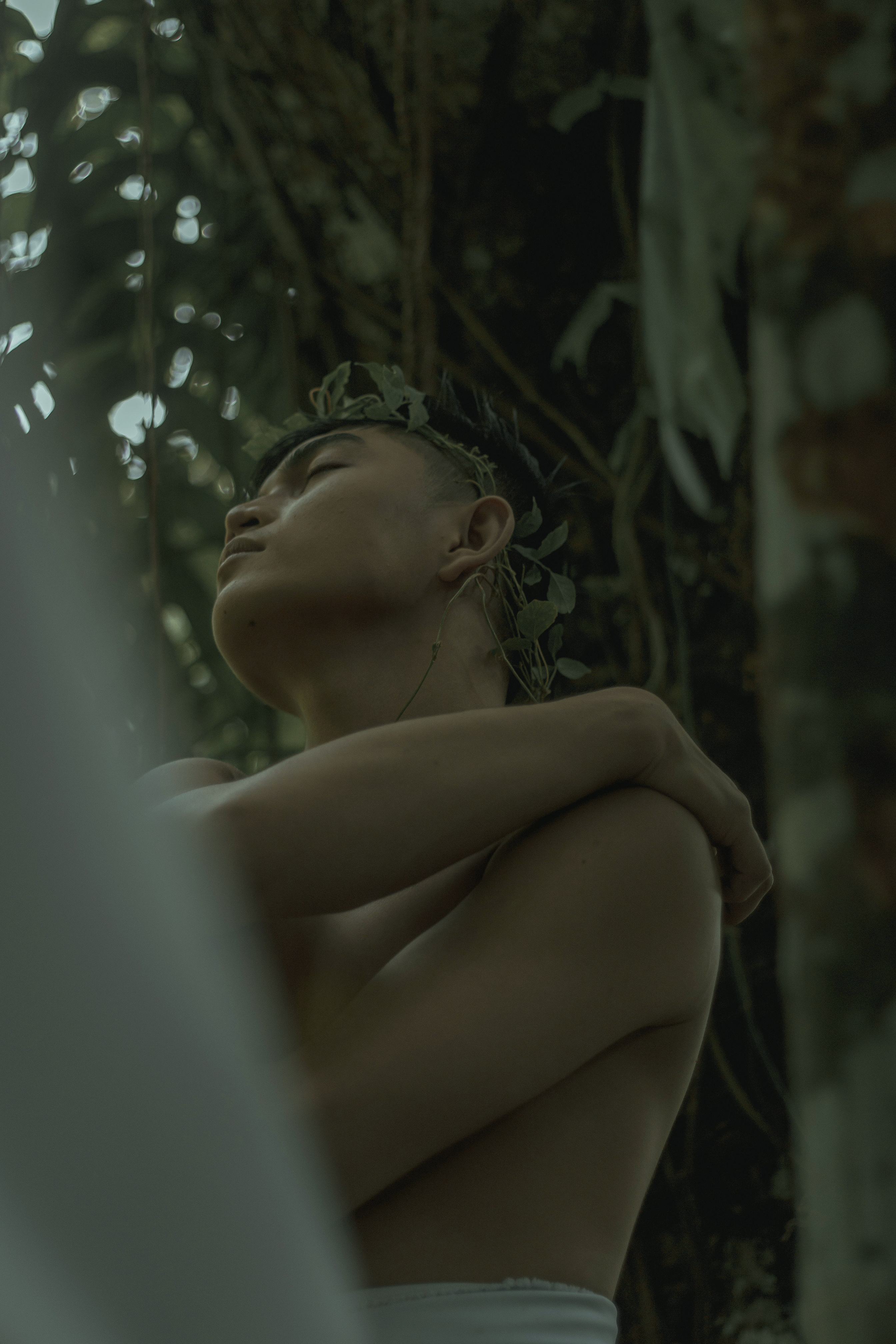 From below of slim dreamy male in wreath touching shoulder in tropical forest on gloomy day