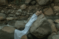 Male in long white sheet resting on heavy rough stone near splashing stream