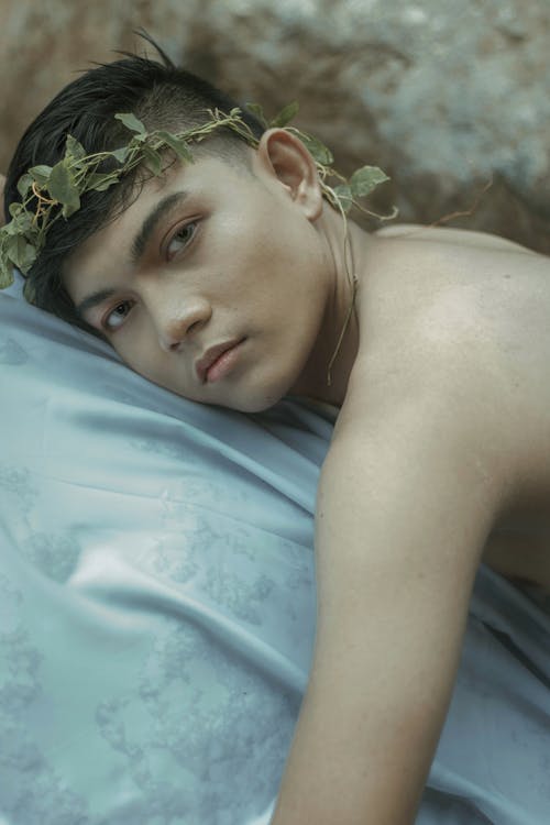 Side view of young Asian man in green wreath on head and bared shoulders lying on stone covered with white cloth and looking at camera