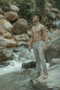 Full body of young ethnic barefoot male looking away and standing on rough stone near swift stream