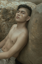Ethnic male with stylish haircut and smear of foundation cream on face looking at camera and resting near heavy rocky stone