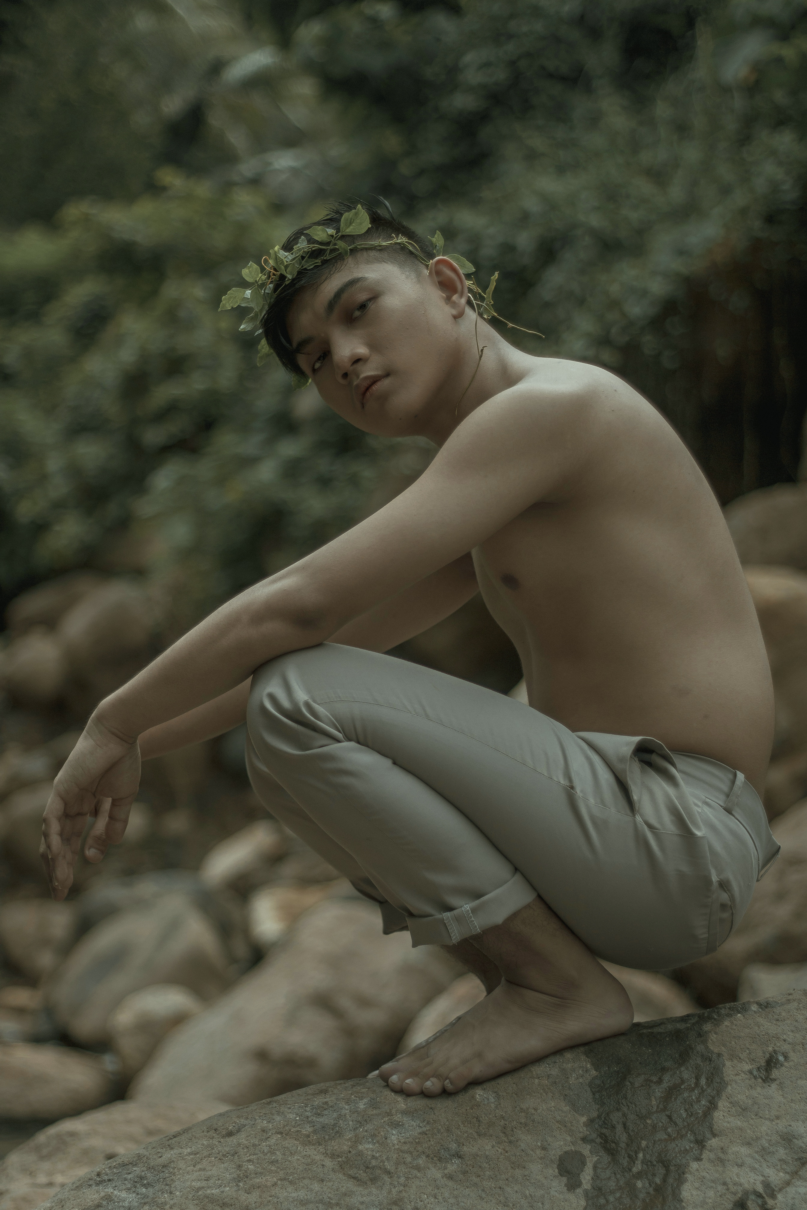 白天，一名头戴叶子花环、裸露上身的年轻民族男子蹲在自然中的石头上