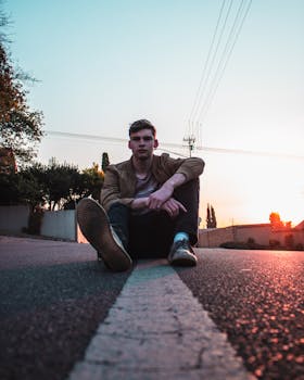 A young man in casual attire sits on a road at sunset, showcasing urban fashion and laid-back style.