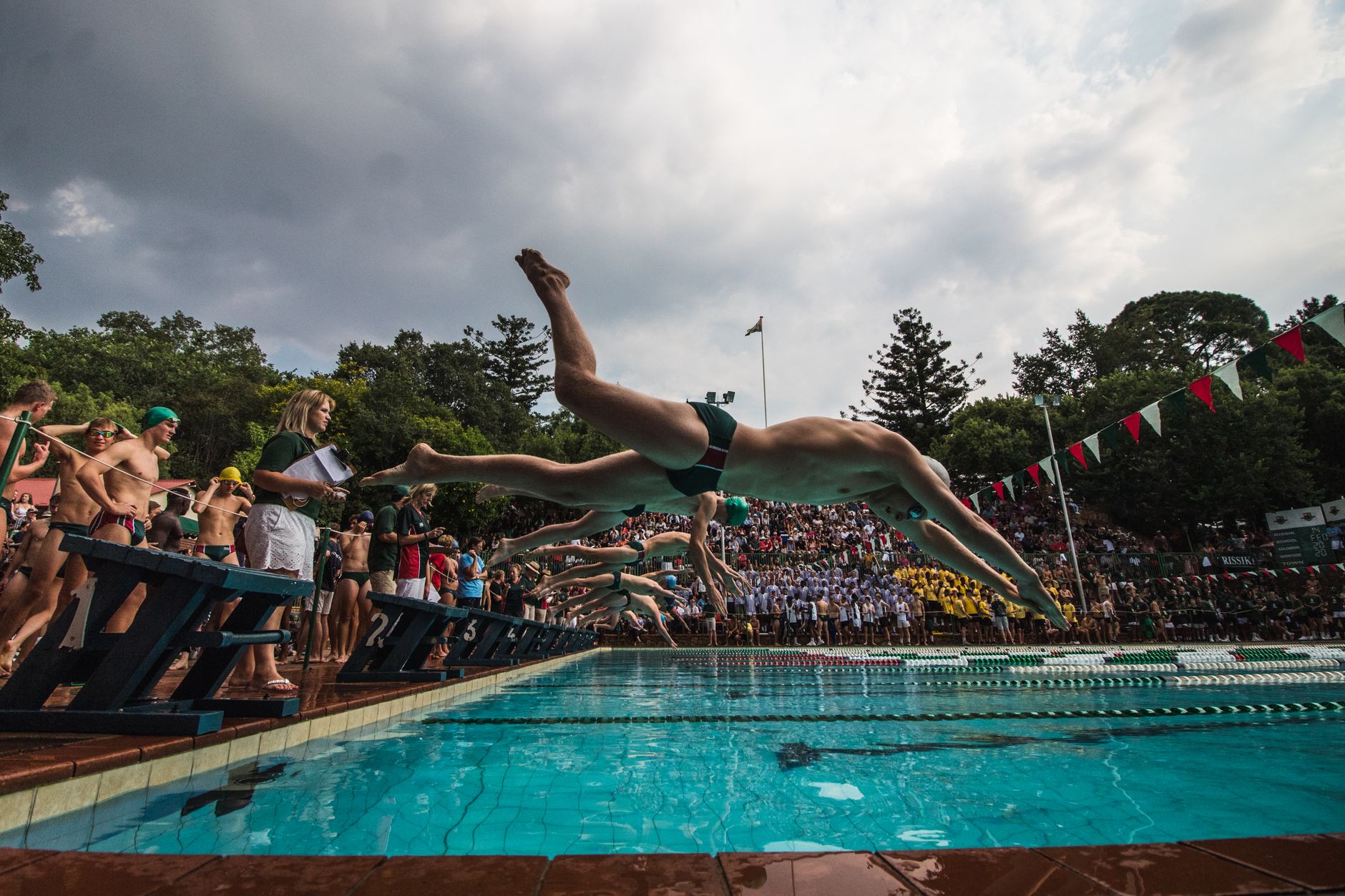 Swimmers in action diving into an outdoor pool during a competitive event with a crowd watching.