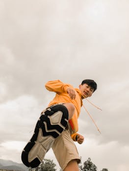 Low angle shot of a young man posing with an outstretched foot against an overcast sky, showing motion and energy.