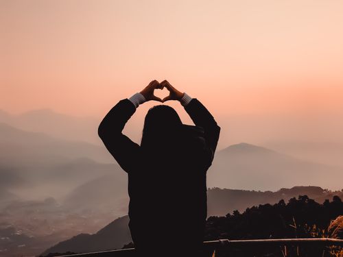 Free A woman makes a heart shape with her hands, silhouetted against a mountain sunset. Stock Photo
