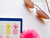 笔记本上放着一副太阳镜、一朵粉色花朵和一个色彩缤纷的菠萝，旁边还有一本时尚的公寓杂志