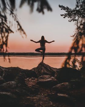 Silhouette of a woman practicing yoga on rocks by the water at sunset, creating a serene outdoor scene.