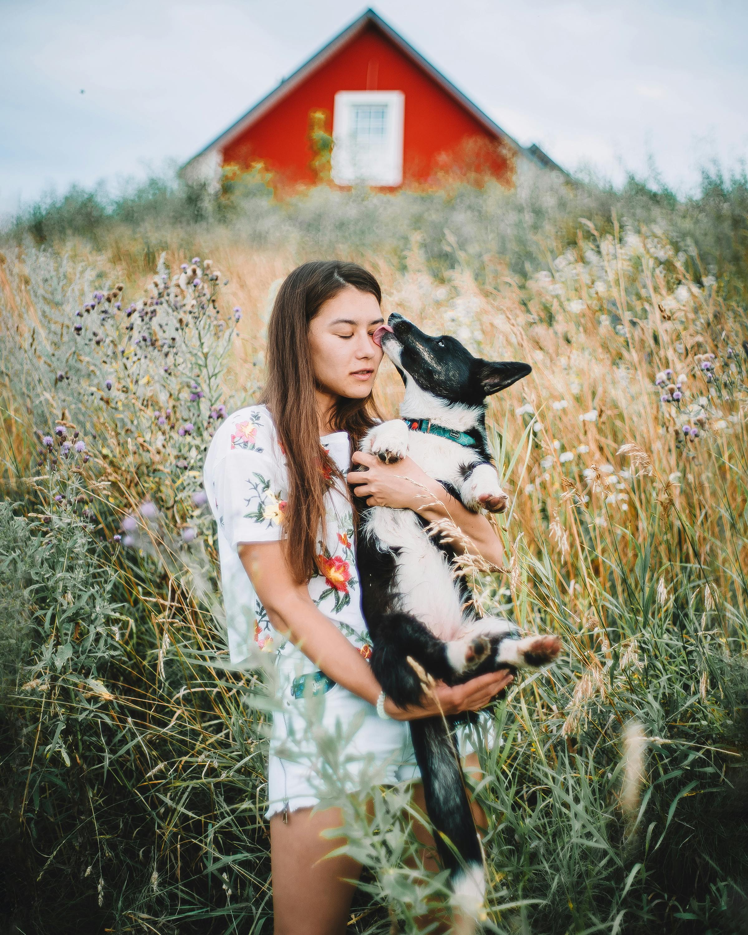 slim woman holding dog in meadow by house under grey sky