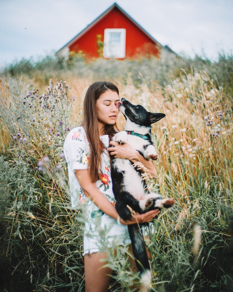 Slim Woman Holding Dog In Meadow By House Under Grey Sky