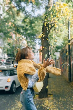 穿着毛衣的女人在城市人行道上扔秋叶，微笑着捕捉秋天的喜悦