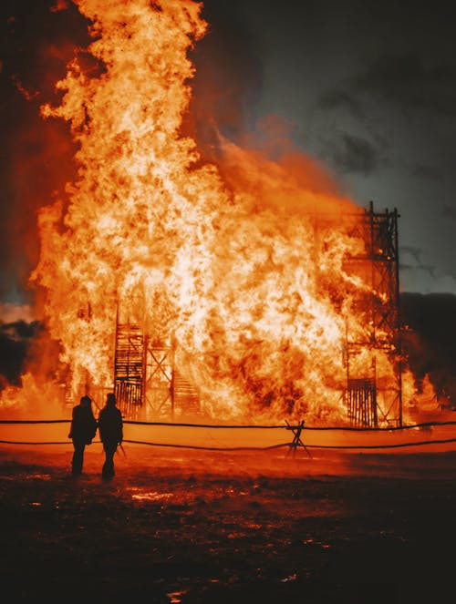 Silhouette of faceless people standing next to bright orange burning construction against dark sky in evening