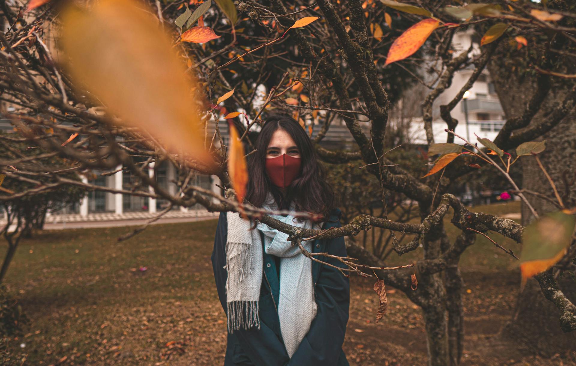 Woman in autumn park wearing facemask amidst falling leaves, embracing seasonal vibes and safety.