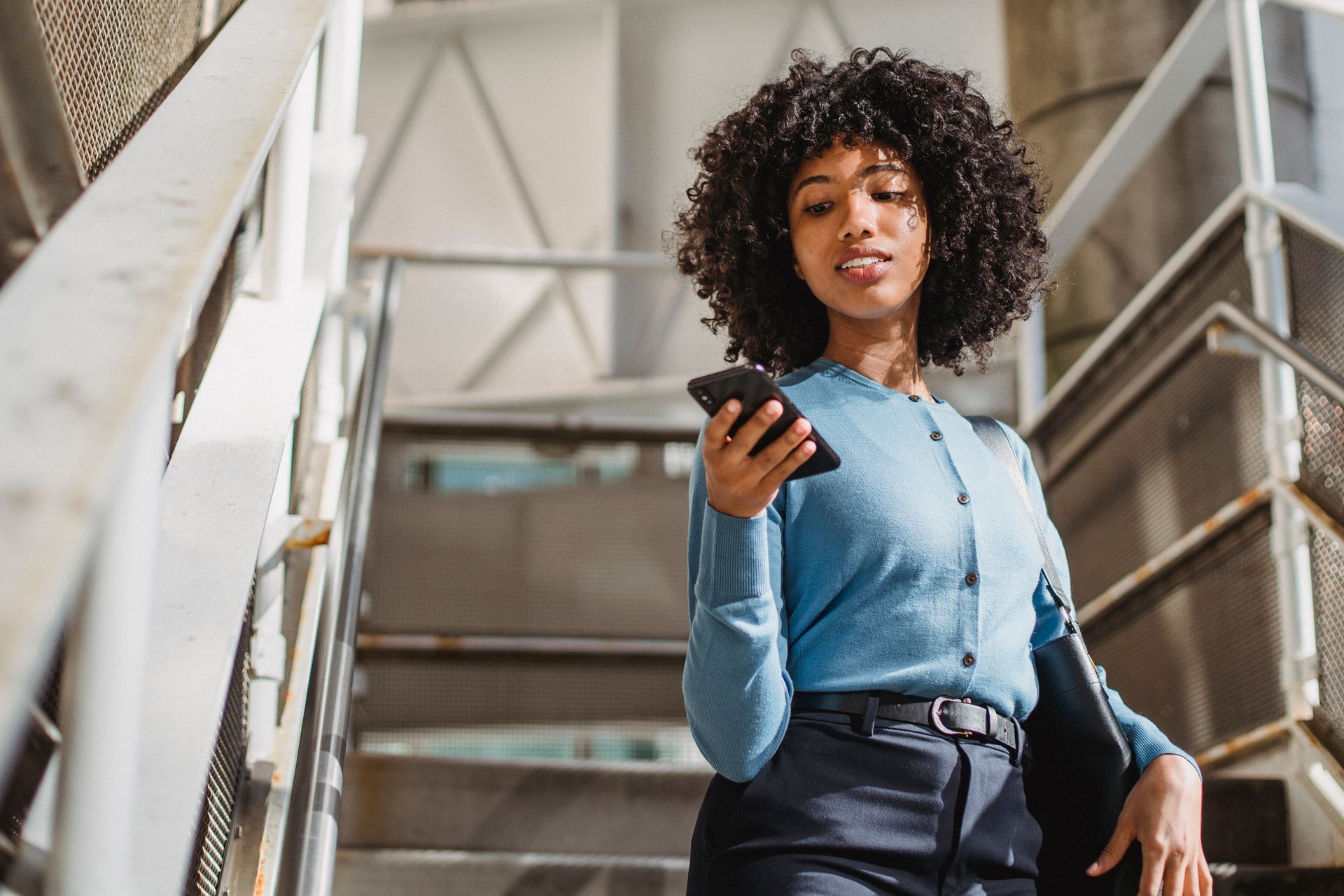From below of cheerful elegant African American female texting message on cellphone while standing on stairs with railings on street