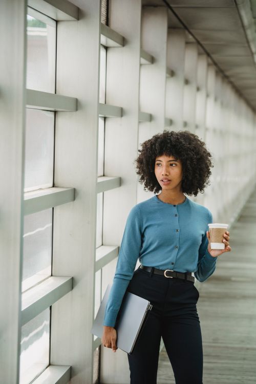 Free Thoughtful African American female with paper cup of hot beverage standing with laptop and looking out window during coffee break Stock Photo