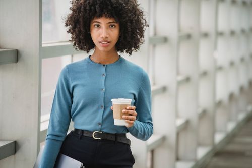 Free Content African American female freelancer with Afro hairstyle and takeaway coffee looking at camera while standing with laptop near window on blurred background Stock Photo