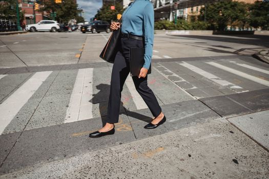Unrecognizable female in casual wear with black bag walking on pedestrian crossing on sunny street in city with buildings on background