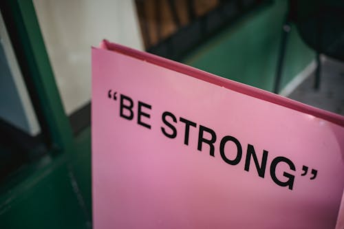 From above of pink signboard with black motivational phrase Be strong on street near green facade of building in city with blurred background